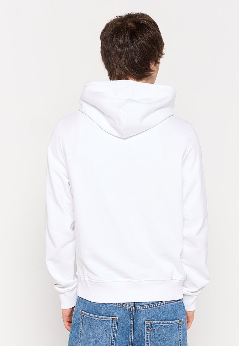 Persona con capelli corti castani che indossa una felpa bianca e jeans di denim blu, rivolta di spalle su uno sfondo bianco uniforme.