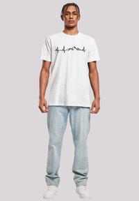 White cotton t-shirt with short sleeves featuring a black mountain and heartbeat line graphic. Paired with light blue denim jeans and white sneakers.