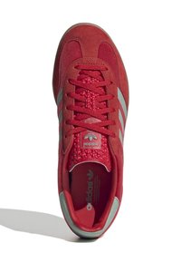Chaussure de sport rouge avec une tige en daim, un motif à trois bandes grises, des lacets rouges texturés et un col rembourré avec des détails de logo.