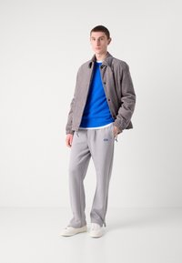 Chaqueta de pana gris, suéter de punto azul, pantalones grises con cordón y cremalleras, y zapatillas blancas. Modelo de pie contra un fondo blanco.