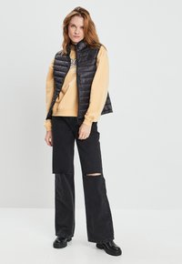 Zwarte puffervest met een hoge kraag, gedragen over een lichtgele sweatshirt. Gepaard met gescheurde zwarte jeans en robuuste zwarte laarzen.