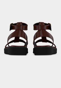 Καφέ δερμάτινα σανδάλια με πολλαπλούς ιμάντες και κούμπωμα με αγκράφα. Μαύρη υφή σόλας με οδοντωτά άκρα για καλύτερη πρόσφυση. Εμφανίζεται η πίσω όψη.