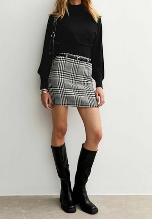 Femme portant un haut noir à manches longues, une mini-jupe à carreaux noir et blanc avec ceinture, des bottes noires montant jusqu'aux genoux, et un sac à épaule noir.