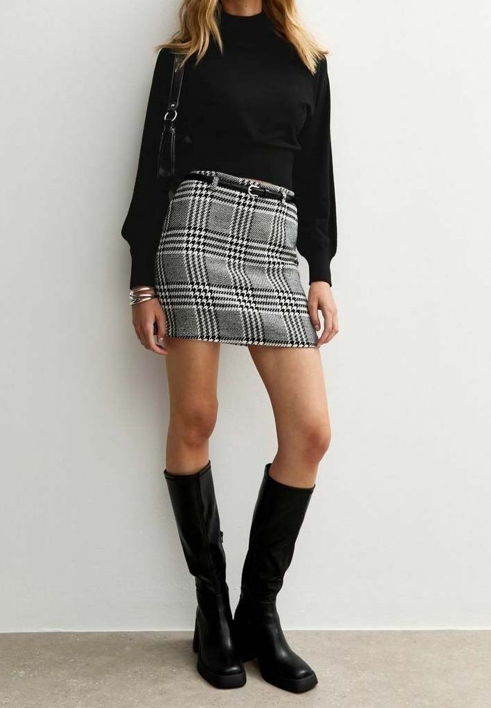 Femme portant un haut noir à manches longues, une mini-jupe à carreaux noir et blanc avec ceinture, des bottes noires montant jusqu'aux genoux, et un sac à épaule noir.