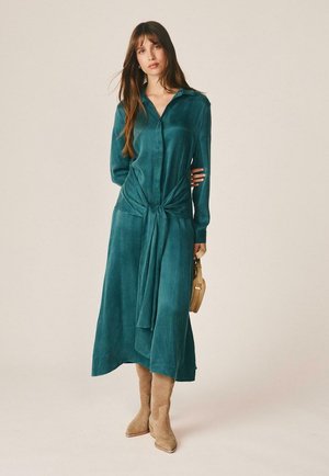 Mujer con pelo largo vistiendo un vestido midi de manga larga color teal atado a la cintura, botas de ante color tostado y sosteniendo un pequeño bolso beige.