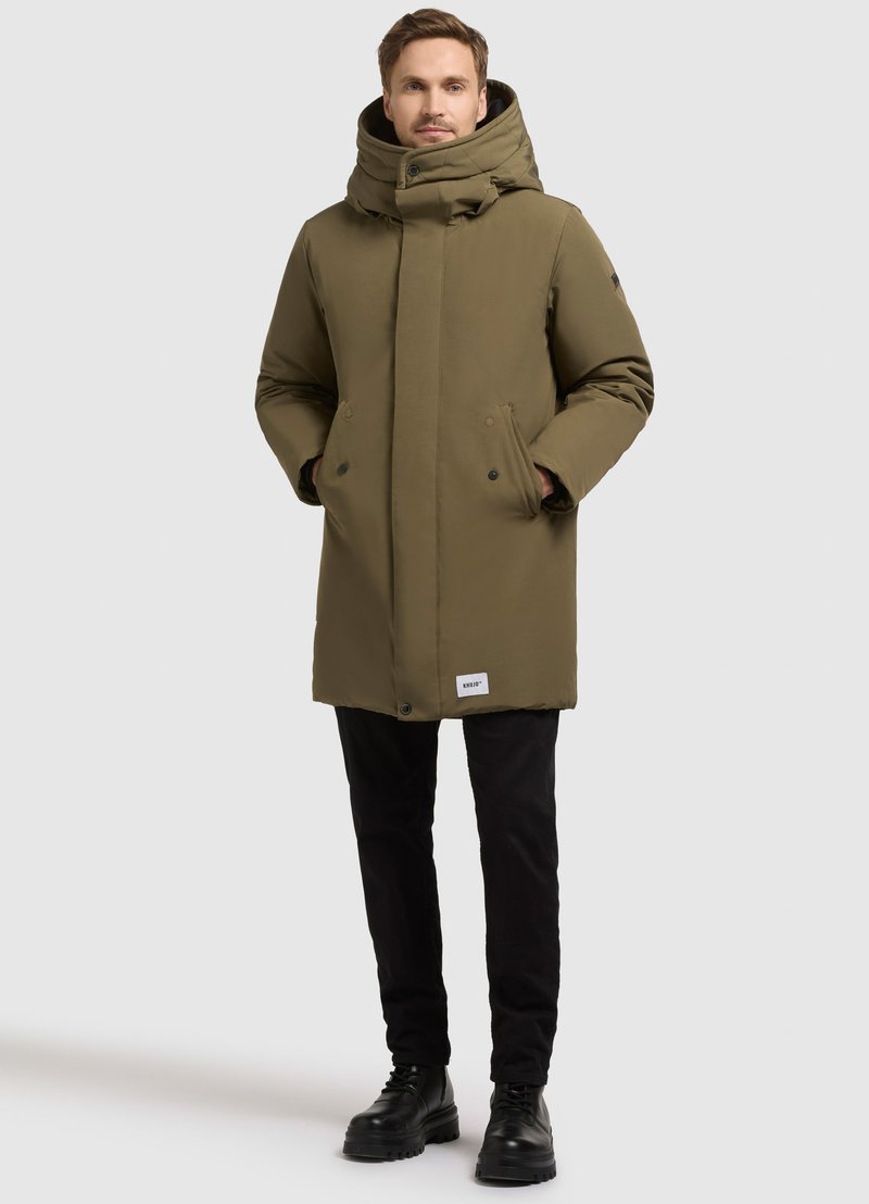 Kirkas oliivivihreä eristetty parka, jossa on korkea kaulus, kaksi etutaskua, nepparilukot ja mustat yksityiskohdat; yhdistetty mustien housujen ja kenkien kanssa.