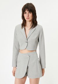 Blazer grigio chiaro corto con chiusura a singolo bottone e tasca frontale, abbinato a pantaloni corti a vita alta coordinati con zip laterale.