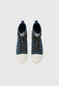 Aukštos sportinės bateliai, pagaminti iš džinsinio audinio, su baltu guminiu noskos dangteliu ir juodomis raištelėmis, ant kurių yra prekės ženklo logo. Tekstūruota mėlyna medžiaga su nusidėvėjusiomis detalėmis.