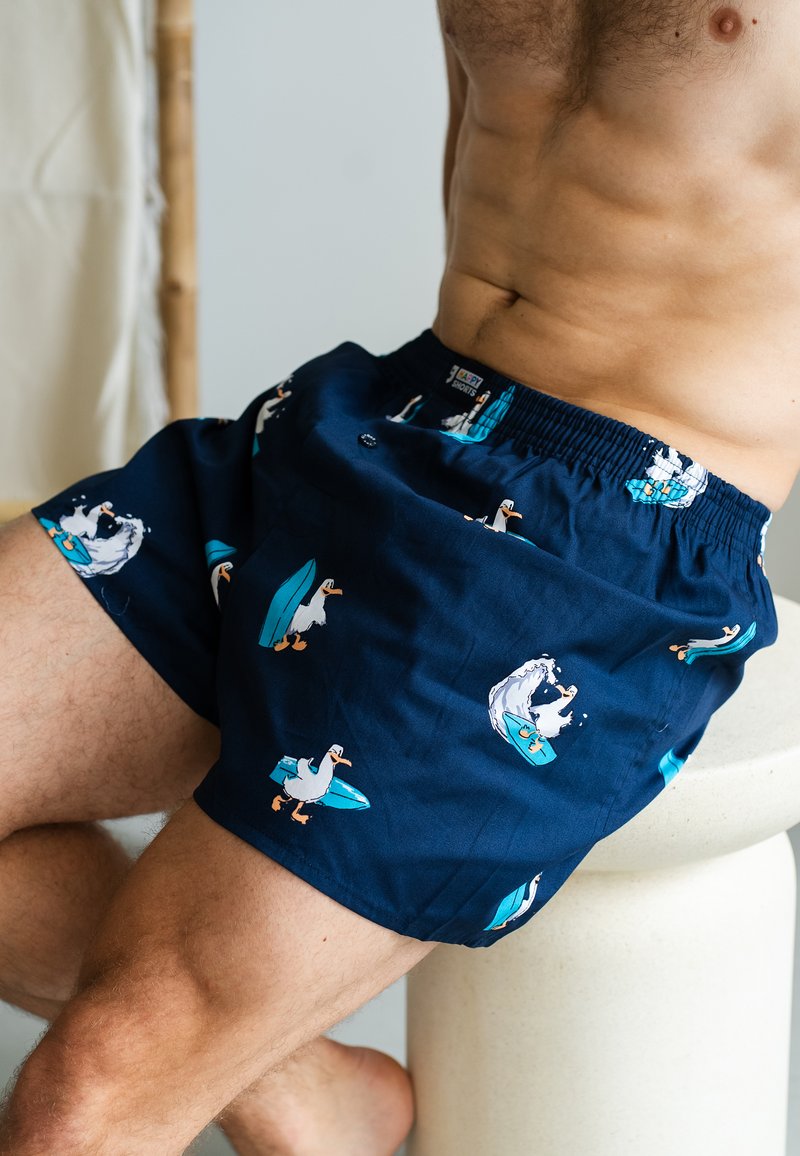 Marineblaue Schwimmshorts mit einem Muster von Surfdiensten. Elastischer Bund mit Knopfdetail und weichem, leichtem Stoff für zusätzlichen Komfort.