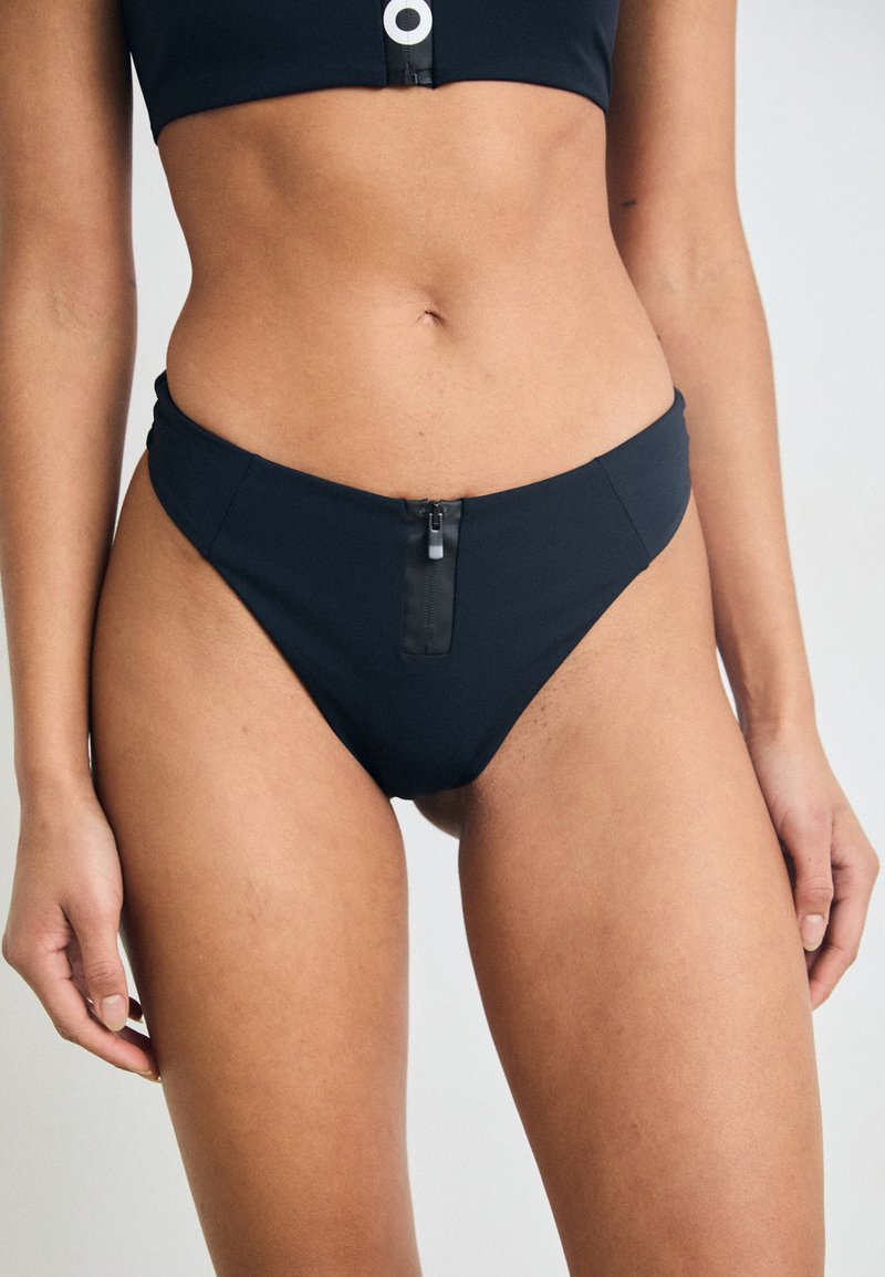 Melnās bikini apakšdaļas, kas izgatavota no gludas auduma, ar priekšējo rāvējslēdzēju, vidēju pārklājumu un tīru, minimālistisku dizainu.
