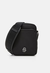 Sac crossbody noir en tissu texturé avec fermeture éclair. Présente un accent logo et une sangle ajustable à motif. Design compact et carré.