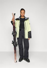 Die Skiausrüstung besteht aus einer hellgrünen wasserdichten Jacke mit schwarzen Akzenten, kombiniert mit schwarzen Schneehosen. Die Person hält einen braunen Ski mit Bindungen.
