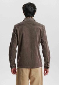 Cămașă de culoare maro din corduroy, cu mâneci lungi, manșoane cu nasturi și guler. Prezintă un umăr din spate și o croială lejeră. Purtați cu pantaloni de culoare deschisă.