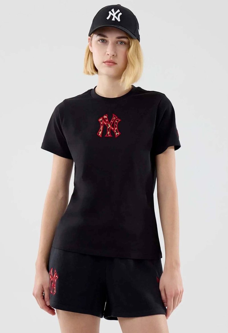 Jeune femme portant une casquette noire, un T-shirt noir et un short noir, tous avec des logos NY rouges et blancs, debout sur un fond uni.