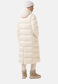 Vrouw in een lange crèmekleurige pufferjas met capuchon, crèmekleurige broek, beige laarzen met bontvoering en een witte gebreide beanie, staand met haar zij naar de camera gericht.