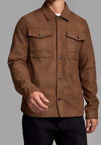 Homme portant une veste en daim marron à boutons avec des poches poitrine à rabat et un pantalon noir, se tenant contre un fond gris uni.