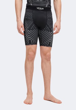 Homme portant un short de compression noir et gris à motif géométrique avec une ceinture à logo « Venum », debout pieds nus devant un fond neutre.