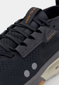 Chaussure de sport noire avec une tige perforée, des superpositions texturées et des accents réfléchissants. Présente les inscriptions "ALL NIGHT" et "NIKE TRAIL" en orange.