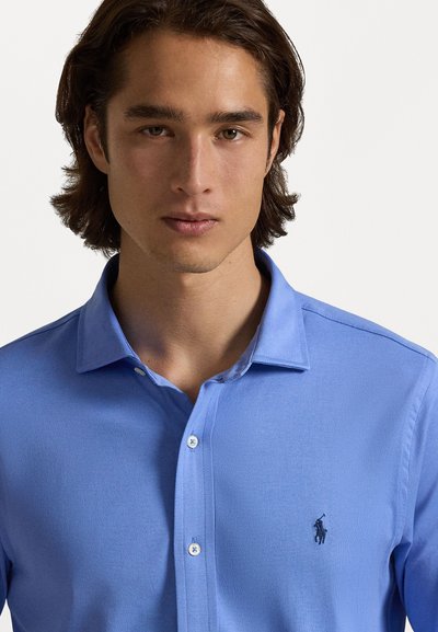 Camisa polo de cor azul claro com colarinho, feita de um tecido suave, apresentando um fecho frontal com botões e um logótipo bordado no lado esquerdo do peito. Textura suave.