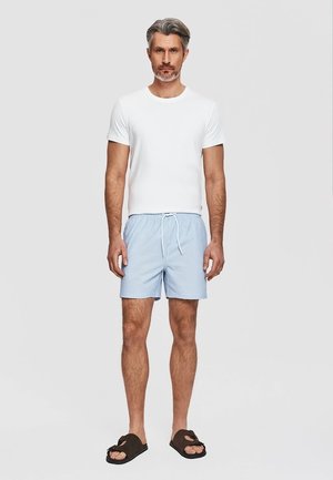 Mężczyzna stoi, ubrany w białą koszulę z krótkim rękawem, jasnoniebieskie krótkie spodenki ze sznurkiem i ciemne sandały na jednolitym jasnym tle.