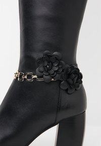 Czarny skórzany botek za kostkę z ozdobą w postaci złotego łańcuszka i teksturowanymi kwiatowymi detalami, na masywnym obcasie i gładkiej powierzchni.