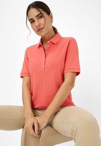 Polo shirt corallo con colletto e placket a cinque bottoni. Maniche corte e texture morbida. Indossato con pantaloni beige. Design semplice, senza modelli visibili.