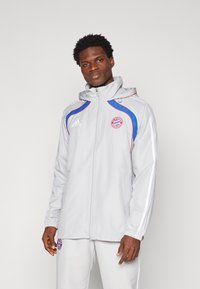 Lichtgrijze sportjack met een capuchon, voorzien van blauwe en rode accenten, een ritsluiting en een logo op de borst. Inclusief elastische manchetten en zijzakken.