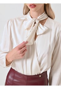 Blusa crema con dettaglio collo a fiocco, tessuto morbido, maniche lunghe e polsini arricciati, abbinata a una gonna in finta pelle bordeaux a vita alta.