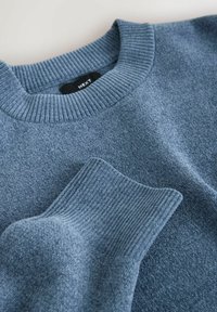 Pulover tricotat albastru cu guler și manșete cu nervuri, așezat plat pentru a evidenția textura și detaliile în prim-plan ale materialului și coaserii.