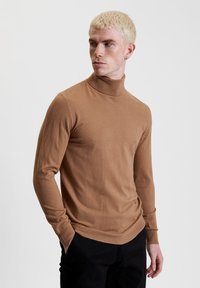 Maglione a collo alto marrone realizzato in tessuto morbido, slim fit, maniche lunghe, con collo e orlo a coste, indossato con pantaloni neri.