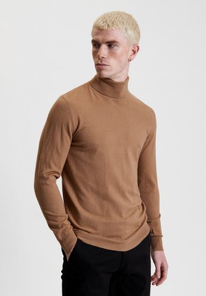 Bruine coltrui gemaakt van zachte stof, slim fit, lange mouwen, met een geribde hals- en onderrand, gedragen met zwarte broeken.