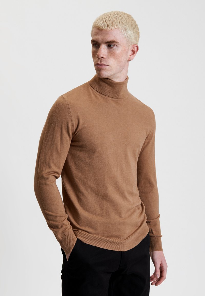 Maglione a collo alto marrone realizzato in tessuto morbido, slim fit, maniche lunghe, con collo e orlo a coste, indossato con pantaloni neri.