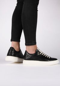 Černé kožené tenisky s bílou gumovou podrážkou, kulatým špičatým designem, krémovými tkaničkami a decentním bočním detailem. Vhodné pro volný čas.