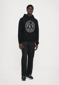 Must mustaga, millel on ees suur ümmargune valge graafiline logo, millel on kirjas "Versace Jeans Couture", kombineeritud tumedate cargo-pükste ja tennikega.
