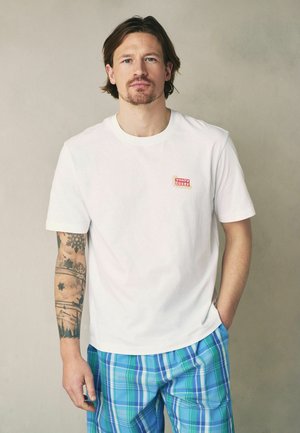 Mężczyzna z tatuażem na ramieniu, ubrany w białą koszulkę "Tony's Diner" i niebiesko-zielone, kratkowane szorty, stojący na jednolitym beżowym tle.