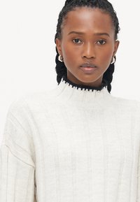 Knit sweater in light cream with vertical ribbed texture, high collar with black trim, and relaxed fit sleeves. Silver hoop earrings visible.