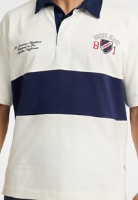 Polo shirt in cream and navy striped design. Features a collar, short sleeves, embroidered logo and text on the left chest. Cotton blend material.