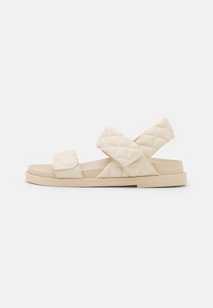 Beige flache Sandale mit gesteppte Stoffriemen, einer über den Zehen und einer, die den Knöchel kreuzt, auf einfarbig hellem Hintergrund.
