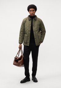 Quiltet grøn jakke med brun krave, to lommer, sort turtleneck, sorte bukser og brun lædertaske, båret med sorte sko.