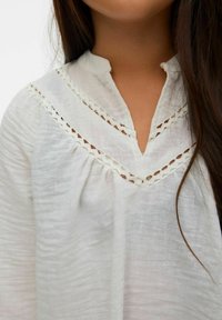 Camicia bianca con tessuto strutturato, caratterizzata da scollo a V e bordo decorativo in pizzo. Design plissettato con vestibilità ampia, morbida e leggera.