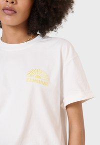 Weißes Baumwoll-T-Shirt mit gelbem, besticktem Sonnenmotiv und dem Wort "SUNSHINE", kurzen Ärmeln und umgeschlagenen Ärmeln. Glatte Textur.