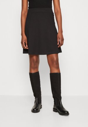 Woman wearing a black knee-length skirt and black knee-high leather boots standing against a white background.