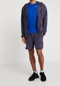 Men's activewear: navy blue moisture-wicking short-sleeve shirt, gray shorts with perforated design, lightweight jacket, black sneakers.