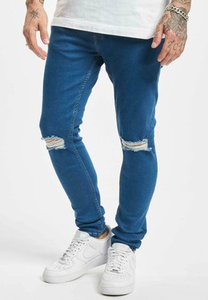 Mies, joka pukeutuu revittyihin sinisiin skinny-farkkuihin, valkoisiin tennareihin ja valkoiseen paita, esittelee tatuoituja käsiä ja hopeaketjulahjakkeita.