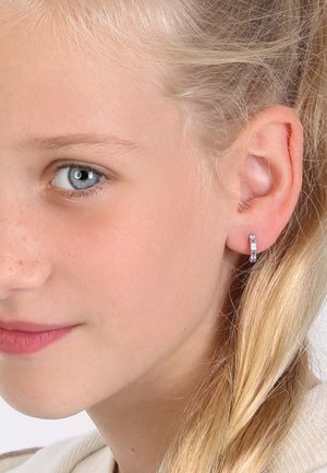 Silberne Creolen mit kleinen klaren Edelsteinen, die ein elegantes Design zeigen. Das Haar ist zu einem Zopf neben dem Ohr frisiert.