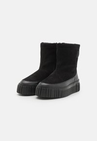Botas negras hasta el tobillo que combinan materiales de ante y cuero suave, con una suela de plataforma gruesa y un detalle texturizado en el borde inferior.