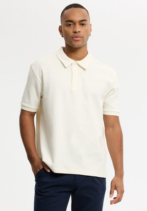 Joven con el pelo corto, vestido con una camisa polo blanca de manga corta y pantalones oscuros, de pie con una mano en el bolsillo sobre fondo blanco.