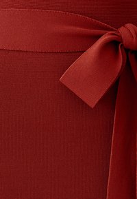 Tela roja texturizada con una cinturilla ancha, que presenta un detalle de lazo atado en un lado. El material parece elástico y suave.
