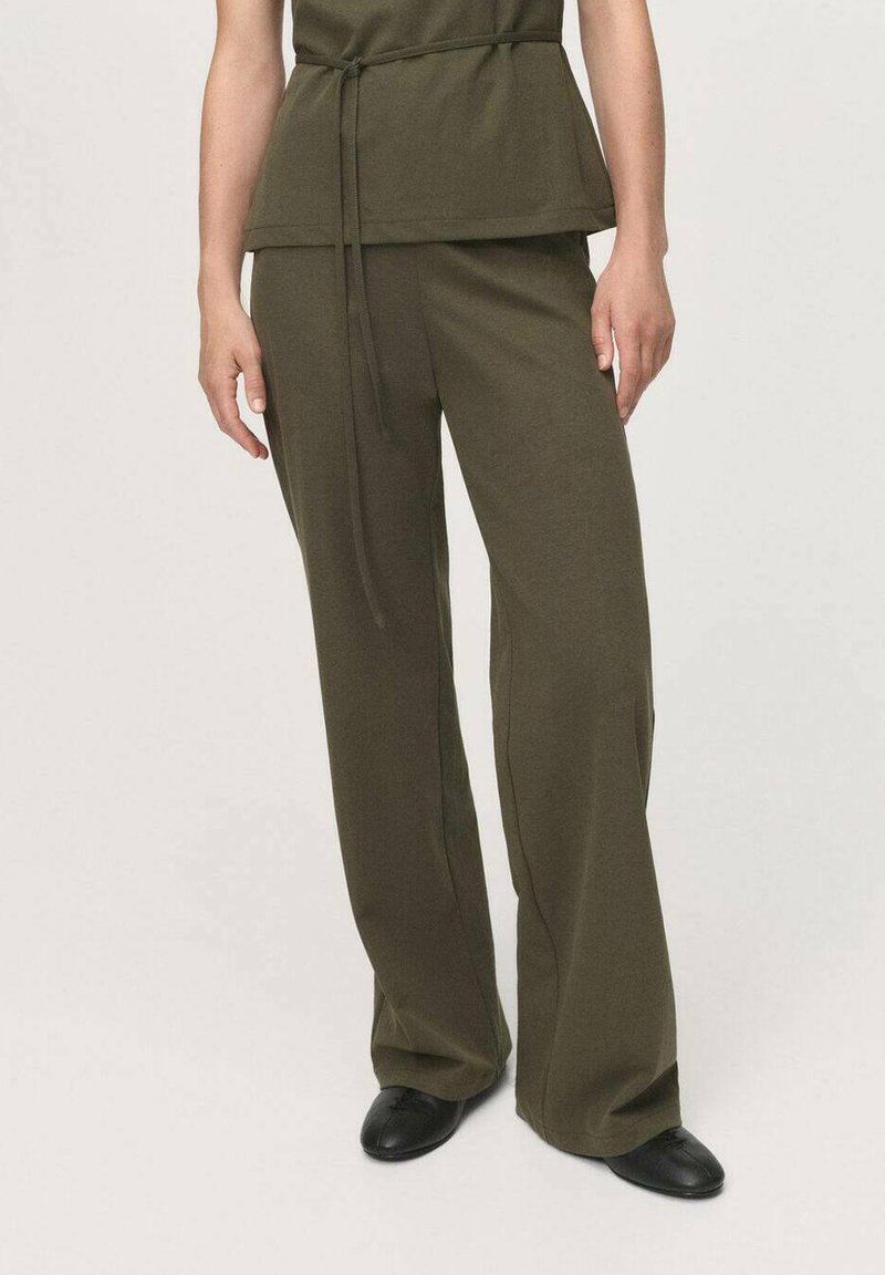 Olijfgroene peplumtop met een ceintuur in de taille en wijde broekspijpen, gemaakt van een zachte stof. Zwarte platte schoenen maken de outfit af.