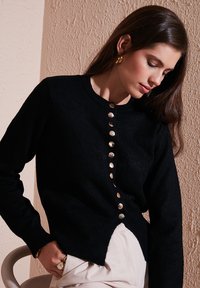 Svart stickad cardigan med rund halsringning, prydd med en rad metallknappar fram och en något längre bakre nederkant.